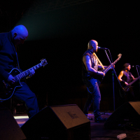 Three musicians perform on a dimly lit stage at the 2012 Kilkim Žaibu festival. The guitarist on the left, dressed in black, plays energetically. The central figure, the lead singer and guitarist, sings into a microphone. The bassist on the right is playing with focus. Amplifiers and stage equipment surround them.