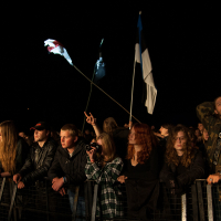 A crowd of people stands behind a barricade at a nighttime outdoor event or concert. Some attendees are holding flags, including a dark and white one. A person wears a camouflage jacket while others are bundled in jackets and hoodies. A small camera captures the scene at Kilkim Žaibu 2012 festival.