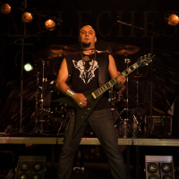 A musician in a black sleeveless shirt with a white graphic plays a black, V-shaped electric guitar on a dimly lit stage at the 2012 Kilkim Žaibu festival. He wears dark pants and stands in front of a drum set. Overhead stage lights illuminate him as he performs, with part of the band banner in the background.