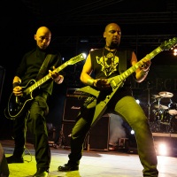 Two bald musicians play electric guitars on stage during a live performance at the 2012 Kilkim Žaibu music festival. One wears a sleeveless top with a skull design and stands in a wide stance. The other is dressed all in black with a collared shirt. Drum equipment, speakers, and stage lighting complete the scene.