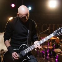 A bald musician wearing a black shirt and wristband is intensely playing an electric guitar on stage under bright lights at Kilkim Žaibu 2012. A drum set and another band member are visible in the background. The stage is illuminated with colorful lights, creating a vibrant atmosphere.