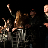 Concertgoers stand behind a metal barricade at an outdoor nighttime event in Lithuania. Several have their arms raised, some with clenched fists. Dressed in dark clothing, the lighting captures their enthusiastic expressions as they engage with the performance, reminiscent of Kilkim Žaibu 2012 festival vibes.