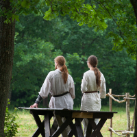 Two people with long hair stand near a wooden bench in a forest clearing. Dressed in medieval-style clothing reminiscent of Kilkim Žaibu 2012, they face away from the camera, towards a sunlit, green background. A tree's branches frame the top of the image, providing partial shade. Wooden structures are visible in the distance.
