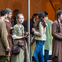 A group of young people dressed in medieval or fantasy-inspired costumes stand under a tent at the 2012 Kilkim Žaibu festival. Two men and a woman in the foreground are engaged in conversation, while the woman on the right holds a wooden bowl and looks contemplative. Modern clothing is visible in the background.