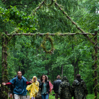 Several people, some wearing raincoats, walk through a makeshift archway made of intertwining branches and greenery on a forest path during the 2012 Kilkim Žaibu festival. It is raining, and the lush surroundings indicate a dense forest. The atmosphere appears to be wet and possibly chilly.