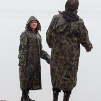 Two people wearing long camouflage raincoats and black boots stand on a sandy beach during Kilkim Žaibu 2012. One person faces the camera with their hood up, while the other has their back turned, looking toward the water. The overcast sky suggests a rainy or misty day.