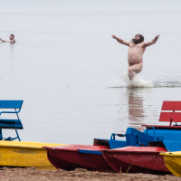 A man joyfully runs out of the water with arms raised at a beach, clearly enjoying the moment. Two other people are seen further in the water, engaged in conversation. In the foreground, brightly painted red, yellow, and blue paddle boats are lined up on the sandy shore—a perfect scene from Kilkim Žaibu 2012.