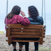 Two people with long hair sit side by side on a wooden swing overlooking a body of water, reminiscent of a serene moment captured at Kilkim Žaibu 2012. The person on the left wears a red jacket and black boots, while the one on the right sports a black leather jacket. They sit closely together, with the person on the left raising their hand to their head.