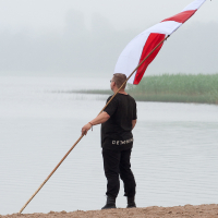 A person standing on a sandy beach holds a large red and white flag mounted on a long pole. The individual, dressed in black with "DETOX" written on the back of their shirt, seems to be part of the 2012 Kilkim Žaibu festival. The background features a foggy lake with blurred vegetation in the distance.