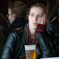 A young person with light brown hair, wearing a studded black leather jacket and a red plaid shirt, rests their head on their hand. They are seated at a table with a plastic cup of beer in front of them at Kilkim Žaibu 2012 festival. Their expression appears contemplative while another person is visible in the background.