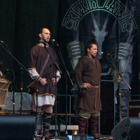 Four men dressed in medieval-style clothing stand on a stage at the 2012 Kilkim Žaibu festival. Two of them play drums while the others sing into microphones. Behind them is a banner featuring a goat skull and the word "Vikivarri" in stylized letters. The dark backdrop emphasizes the performers.