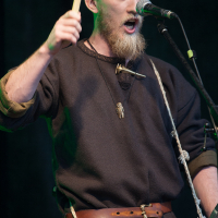 A person with a beard is enthusiastically playing a drum using a mallet at Kilkim Žaibu 2012. They are dressed in dark, medieval-style clothing and are singing into a microphone on a stand. The background is dark, emphasizing the musician performing on stage at the festival.