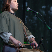 At the 2012 Kilkim Žaibu festival, a person with shoulder-length hair is playing a drum and singing into a microphone on a stand. They are wearing a green tunic with rolled-up sleeves and have a pouch hanging at their side. The image captures an outdoor performance with a dark, blurred background.