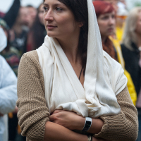 A woman wearing a cream-colored shawl over a brown knit sweater stands with her arms folded in a crowd at the 2012 Kilkim Žaibu festival. She has dark hair and is gazing thoughtfully to the side. The background is blurred, showing other people and some greenery, suggesting an outdoor event.