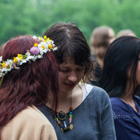 A group of people engaged in conversation outdoors at the 2012 Kilkim Žaibu festival. One person with curly hair wears a necklace and a grey shirt, while another person facing them has long red hair adorned with a floral crown. Other individuals in the background are blurred, with greenery and a wooden post visible.