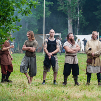 A group of six men dressed in medieval-style clothing stands outdoors on a grassy field with trees and tents in the background at Kilkim Žaibu 2012. They are engaged in conversation and holding various objects. Some have tattoos, and one has a long, braided beard. Their outfits include tunics, belts, and boots.