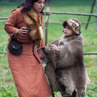 At the 2012 Kilkim Žaibu festival, a woman and child dressed in medieval attire stand on the grass. The woman wears a long brown dress with fur accents and carries a small bag. The child, wrapped in a fur cloak with a matching hat, completes the scene set against wooden fences and tents in the blurred background.