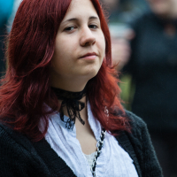 A person with long red hair and fair skin is looking into the camera with a neutral expression. They are wearing a white shirt with a black cardigan and a black choker. The background is blurred with people standing, suggesting an outdoor setting at the 2012 Kilkim Žaibu festival.