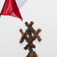 A structure made of hay bales is arranged in a cross-like pattern against a white sky background. In the upper left corner, a red and white flag, reminiscent of the 2012 Kilkim Žaibu festival, is partially visible, fluttering in the wind. The image has a minimalistic and rustic atmosphere.