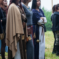 A group of individuals dressed in medieval-style costumes stand outdoors on a grassy field at the 2012 Kilkim Žaibu festival. One person wears a flower crown and a brown cloak, while others don long dresses with sashes and accessories. The background features more people and tall greenery, giving a lively atmosphere.