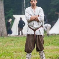 A man stands confidently with arms crossed, wearing medieval-style clothing consisting of a white tunic, brown pants, and wrapped leg garments. Behind him is a forest scene with white tents and other people in similar attire, suggesting a historical reenactment event like Kilkim Žaibu 2012.