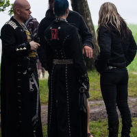 A group of four people stand outdoors near a tree at the Kilkim Žaibu festival. Two are wearing long black coats adorned with metal studs and chains, with one individual having blue hair. Another person is dressed in black with blonde hair, and the fourth, in casual attire, is holding a beverage reminiscent of 2012's carefree vibe.