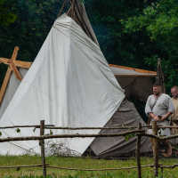 Two men dressed in medieval-style tunics stand outside a large white tent set up in a grassy area with dense green trees in the background. Propped up with wooden poles, the tent and rustic wooden fence create a scene reminiscent of Kilkim Žaibu 2012, adding authenticity to the historical reenactment.