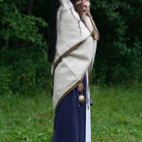 A person stands outdoors on grass with their side profile visible, wrapped in a beige shawl with gold trim. They have long dark hair and are wearing a long blue and white dress. The background consists of dense green foliage, reminiscent of the serene landscape at Kilkim Žaibu 2012. The person appears to be in deep thought or contemplation.