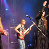 At the 2012 Kilkim Žaibu festival, three musicians perform on stage. From left to right: a woman with long, blonde hair in a red jacket singing, a man with shoulder-length hair in a sleeveless shirt playing a red bass guitar, and a woman with long, dark hair in black clapping near a microphone stand and sheet music. Amplifiers and purple stage lights set
