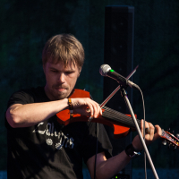 A man with light brown hair is playing a red violin while standing behind a microphone on stage, reminiscent of Kilkim Žaibu 2012. He is wearing a black T-shirt and a bracelet on his right wrist. The background is dark and blurry, with a large black speaker visible behind him. His facial expression shows concentration.