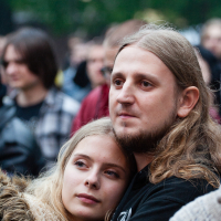 A young woman with long blonde hair rests her head on the shoulder of a young man with long brown hair and a short beard. They appear to be standing in a crowd at an outdoor festival, possibly Kilkim Žaibu 2012. Both have calm expressions. The background is slightly blurred, showing other people and green foliage.