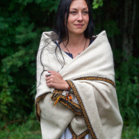 A woman with long black hair stands in front of a lush, green forest background at the 2012 Kilkim Žaibu festival. She is wrapped in a cream-colored blanket adorned with decorative trim. Gazing slightly to the left with a calm expression, her clothing underneath the blanket is white with dark accents.