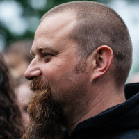 A side profile of a man with a shaved head and a long, bushy beard. He is outdoors, dressed in a black hoodie, smiling as if he's at Kilkim Žaibu music festival. The background is blurred, showing hints of greenery and other people. The lighting suggests it might be overcast or shaded.