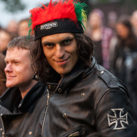 A person with long hair, wearing a headband adorned with red and green feathers and a 5V TURYS label, smiles at the camera at the 2012 Kilkim Žaibu festival. They are dressed in a black leather jacket with a "Chopper" patch on the sleeve. Another person in similar attire is partially visible in the background.
