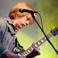 A person with medium-length blond hair plays an electric guitar on stage at Kilkim Žaibu 2012. Dressed in a denim jacket and red checkered shirt, the musician is focused on their performance. A microphone is positioned in front of their face, and a yellow-green background illuminates the scene.