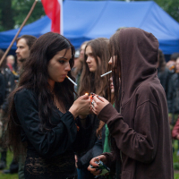 Two women are lighting cigarettes under an overcast sky at the 2012 Kilkim Žaibu festival. One woman with long, dark hair dressed in black ignites a lighter for the other woman in a brown hoodie. A crowd and a flag with white and red horizontal stripes are visible in the blurred background.