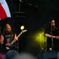 Three musicians perform on an outdoor stage at Kilkim Žaibu 2012. The guitarist on the left wears a black sleeveless shirt and plays an electric guitar. The center musician, clad in dark clothing, passionately plays bagpipes. The drummer in the background is partially obscured as a white and red flag waves in the foreground.