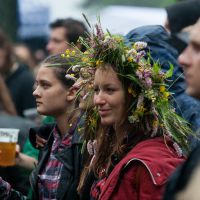 A crowd of people gathered outdoors in 2012, many wearing jackets. In the foreground, a smiling woman wearing a flower crown stands beside another person holding a plastic cup with a beverage. The background shows more individuals at Kilkim Žaibu, some blurred and some focused, indicating various interactions and activities.