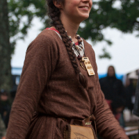A smiling person with long brown hair in braids, wearing a brown medieval-style dress with a belt pouch and a large necklace. The background features trees, blurred figures, and what appears to be an outdoor festival. The person looks happy and engaged, capturing the festive atmosphere of Kilkim Žaibu 2012.