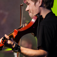 A man with blonde hair plays a red violin on stage at Kilkim Žaibu 2012. He is wearing a black shirt and wristbands, standing in front of a green background. The image captures his side profile, focusing on the instrument, with blurred elements of the band and stage lights in the background.
