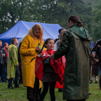 At the 2012 Kilkim Žaibu festival, a group dressed in colorful raincoats stands on a grassy area outdoors in rainy weather. One person in a yellow raincoat holds a bottle, while another in green talks to a child in red at the center. A blue tent is set up in the background, surrounded by trees.