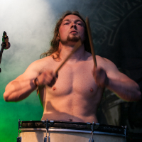 A shirtless man with long hair and a beard is intensely playing a large drum with two sticks. A spotlight shines on him, highlighting his muscular build. The background features an indistinct, dark backdrop with a hint of green lighting, evoking the electrifying atmosphere of Kilkim Žaibu 2012.