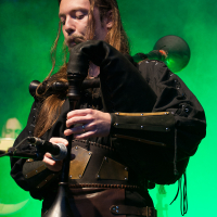 A musician with long hair plays the bagpipes on stage, dressed in medieval-style clothing with a leather vest and metal accents. The background is illuminated with green lighting, creating a dramatic atmosphere reminiscent of Kilkim Žaibu 2012.