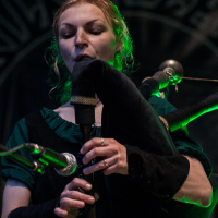 At the Kilkim Žaibu festival in 2012, a woman in a dark green outfit plays the bagpipes with closed eyes, concentrating. The intricate piping is visible. The background features a dark, patterned design illuminated by green light, adding a mysterious ambiance to the scene. She holds the instrument with both hands.
