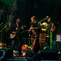 At the 2012 Kilkim Žaibu festival, a band performs on stage with medieval-style instruments. Five members, dressed in medieval-themed costumes, play a variety of instruments including bagpipes, a lute, and drums. The background is dark with green stage lighting and a banner displaying intricate artwork.