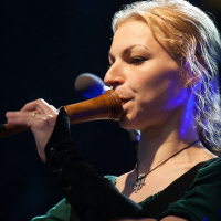 A person with blonde hair is playing a wooden wind instrument on stage at Kilkim Žaibu. They are wearing a green top with long, black fingerless gloves. Blue stage lighting highlights their face, and a microphone is visible in the background, creating an enchanting atmosphere reminiscent of 2012 festival vibes.