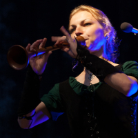 A woman wearing a dark green outfit and arm cuffs plays a wooden flute on stage at Kilkim Žaibu. Blue and purple stage lights illuminate her from the side, creating a dramatic effect. Her blond hair is partially tied back as she stands in front of a dark, blurred background, capturing the essence of 2012.