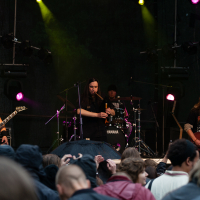 At the 2012 Kilkim Žaibu festival, a rock band performs on an outdoor stage under colored lights, with the lead vocalist at the center holding a microphone. The guitarist is on the left, and a bassist is on the right. A drummer plays in the background. The audience watches, some with rain jackets and umbrellas.