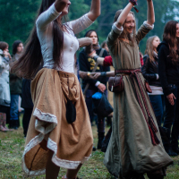 Two women are dancing outdoors at the 2012 festival, dressed in medieval-style clothing. One wears a white blouse with a long beige skirt, and the other wears a green dress with a red belt. They are surrounded by onlookers at what appears to be the Kilkim Žaibu Lithuanian music festival, with trees in the background.