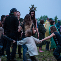 A group of people gathered outdoors are dressed in casual and rain attire. Highlighted in the foreground, children are playing by holding hands and running in a circle. The background features more individuals, trees, a distant treeline, and an overcast sky, suggesting the 2012 Kilkim Žaibu festival.